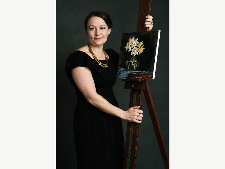 An artist poses for her portrait with one of her paintings as it rests on an easel.