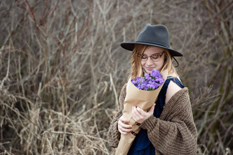 A high school senior sniffs purple flowers while standing in brush and wearing a black hat.