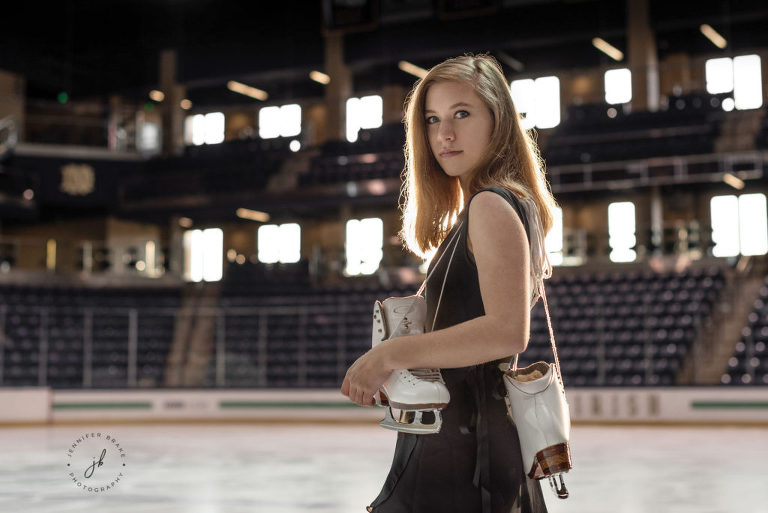 A high school senior figure skater holds her skates over her shoulder as she stands on the ice rink at Compton Family Ice Arena.