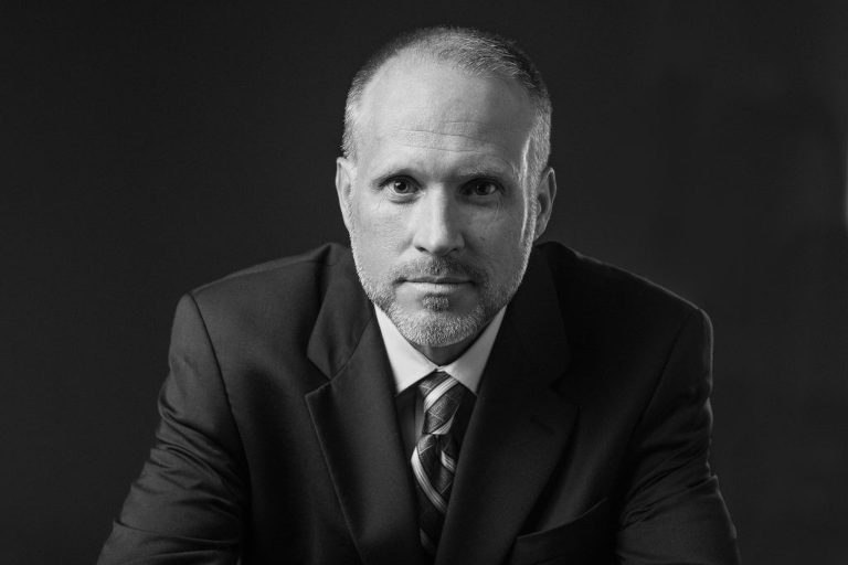 Headshot of a man in a suit and tie leaning forward, listening. Black and white edit.