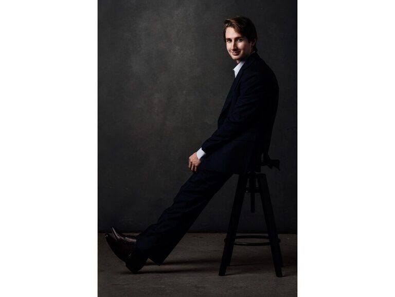 Senior portrait of a guy who leans casually on a stool as he turns toward the viewer.