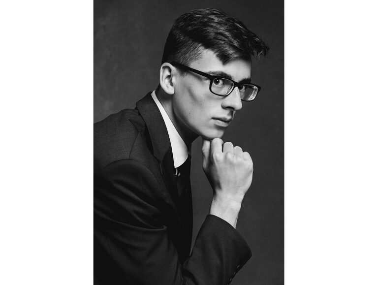 Feature photo: Black and white portrait of a high school senior boy who is wearing a suit and leaning forward with his chin on his thumb and index finger. The light carves out his strong cheekbone and jaw line.